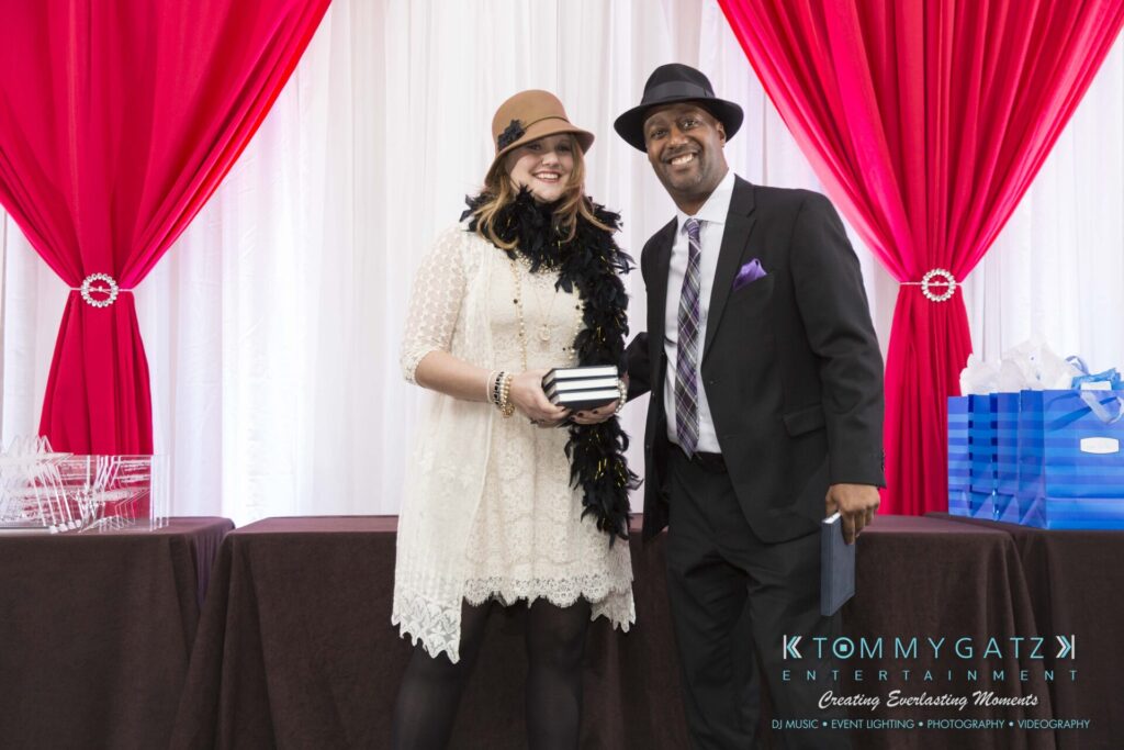 Pillar-Awards-Final-3-24-17-Blog-WM-143 - TOMMY GATZ ENTERTAINMENT A woman and a man in formal attire smile and pose together indoors, standing in front of red and white draped curtains. Several gift bags are on a table nearby.