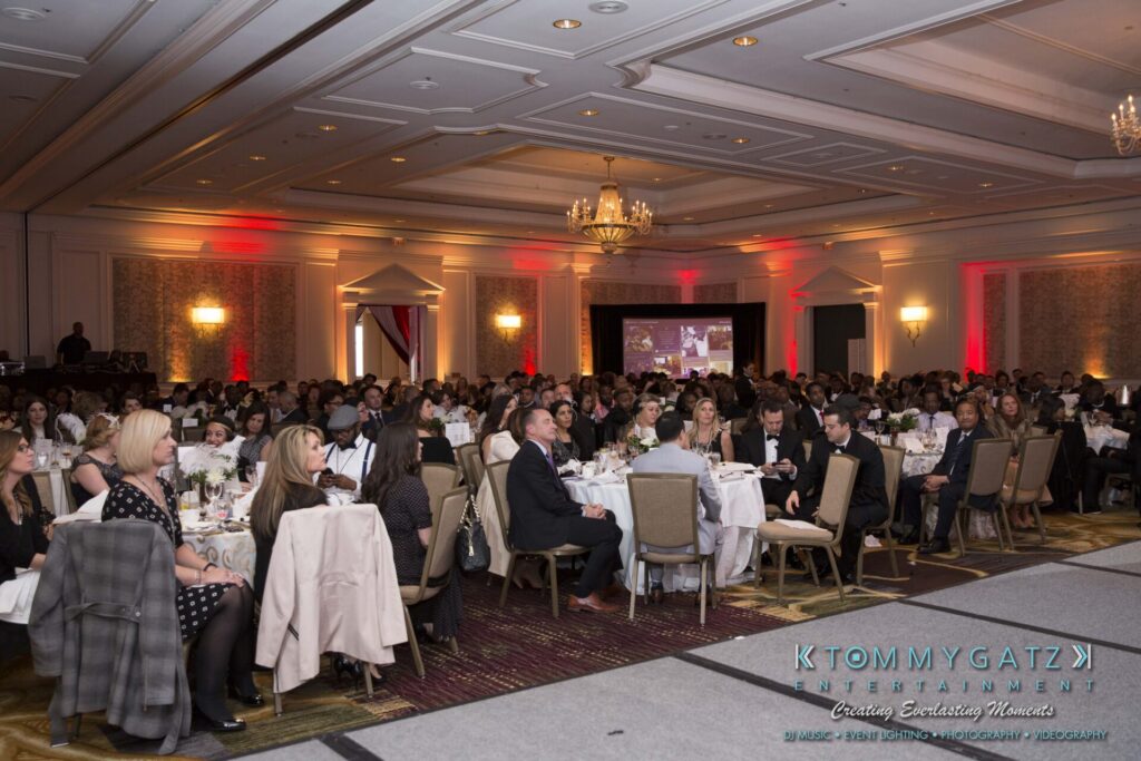 Pillar-Awards-Final-3-24-17-Blog-WM-174 - TOMMY GATZ ENTERTAINMENT A large group of people seated at tables in an elegant banquet hall, attentively facing a stage with a screen displaying a presentation.
