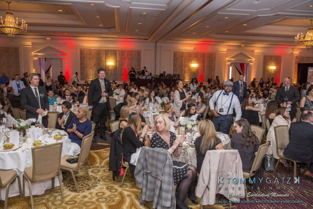 Pillar-Awards-Final-3-24-17-Blog-WM-175 - TOMMY GATZ ENTERTAINMENT A large formal event with many people sitting at round tables in a banquet hall, dressed in business attire, engaged in conversations and activities.