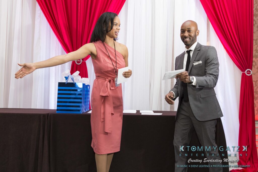 Pillar-Awards-Final-3-24-17-Blog-WM-177 - TOMMY GATZ ENTERTAINMENT A woman in a pink dress gestures towards a man in a gray suit who is smiling and holding an envelope. They stand in front of red and white curtains with a gift bag nearby.