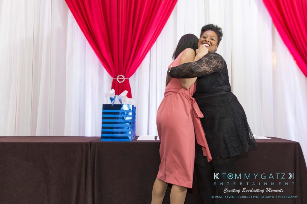 Pillar-Awards-Final-3-24-17-Blog-WM-180 - TOMMY GATZ ENTERTAINMENT Two women embrace joyfully in front of a table with a blue gift bag. A red and white backdrop with "Tommy Gatz Entertainment" branding is visible in the background.