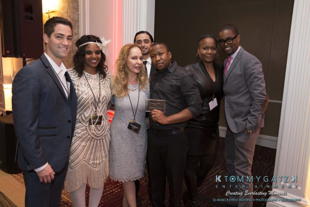 Pillar-Awards-Final-3-24-17-Blog-WM-210 - TOMMY GATZ ENTERTAINMENT A group of seven people standing together indoors, dressed in formal and semi-formal attire, smiling at the camera.