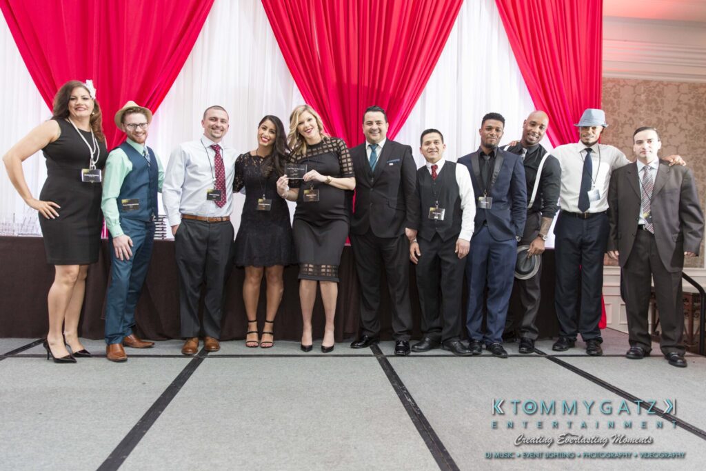 Pillar-Awards-Final-3-24-17-Blog-WM-216 - TOMMY GATZ ENTERTAINMENT A group of people dressed in formal attire stands together on a stage, posing for a photo with a red and white backdrop.