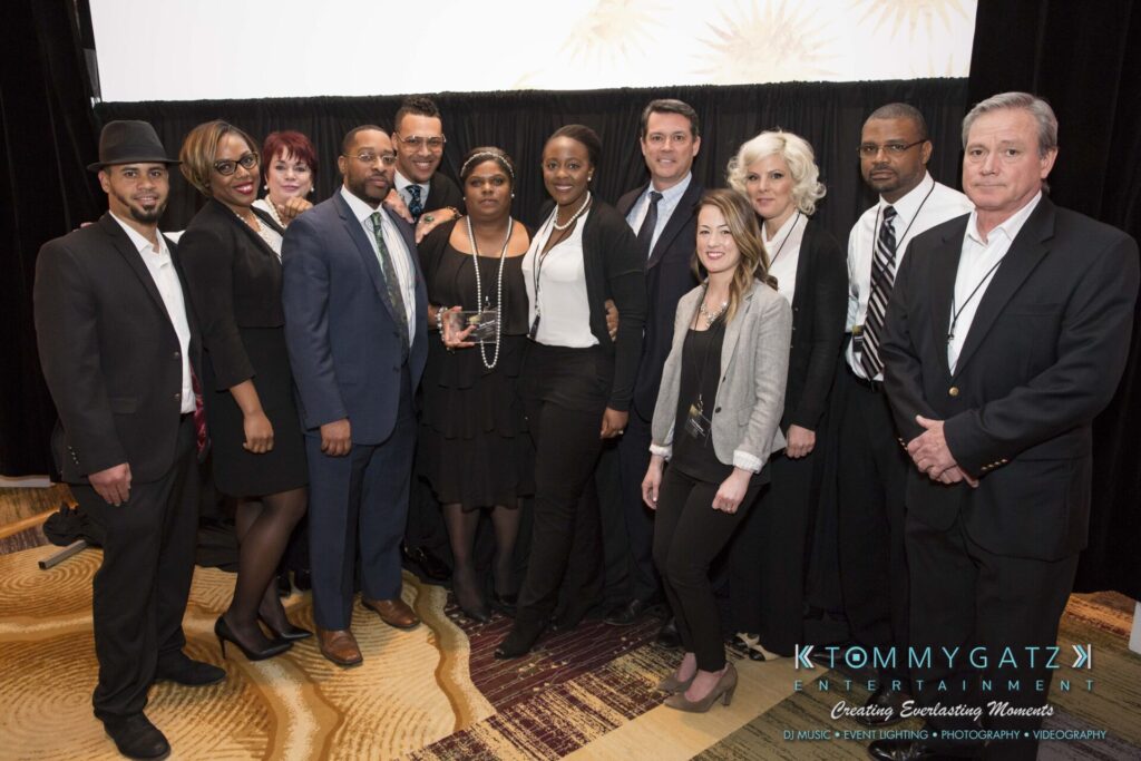 Pillar-Awards-Final-3-24-17-Blog-WM-232 - TOMMY GATZ ENTERTAINMENT A group of 14 people dressed in formal attire posing for a photo in a room with a patterned carpet and dark curtains.