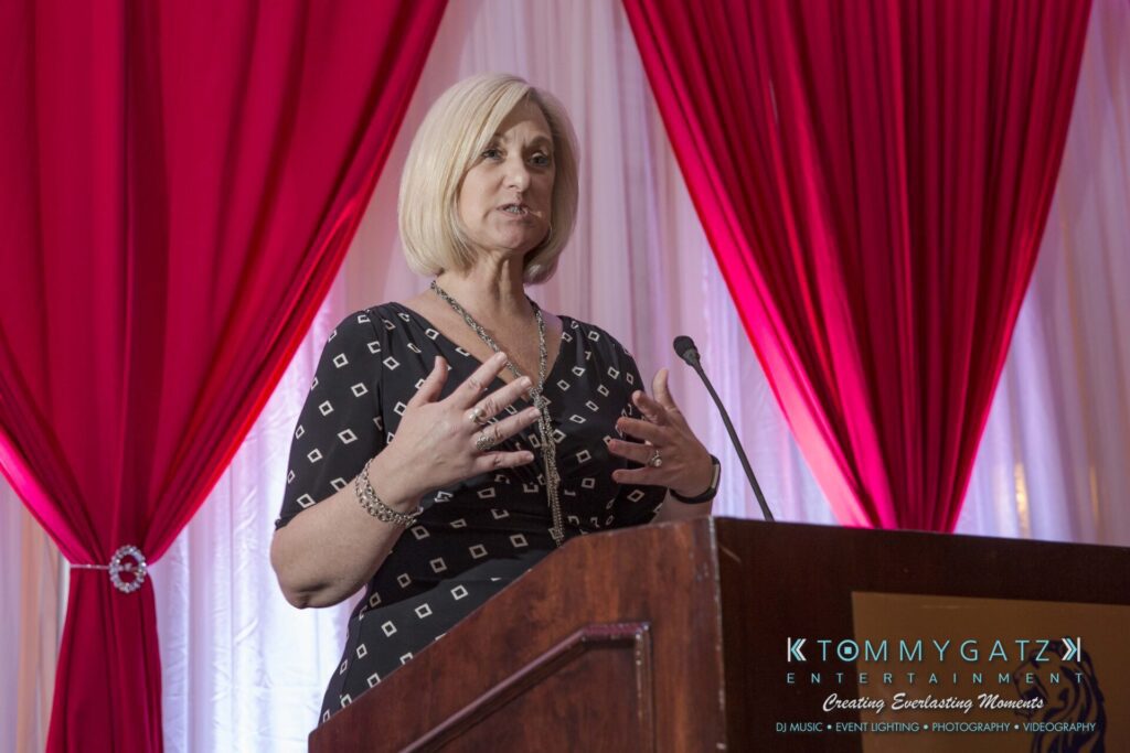 Pillar-Awards-Final-3-24-17-Blog-WM-260 - TOMMY GATZ ENTERTAINMENT A woman speaks at a podium with a microphone, standing in front of red and white draped curtains.