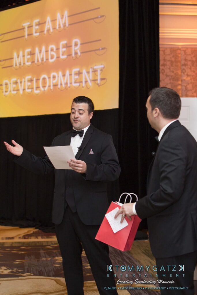 Pillar-Awards-Final-3-24-17-Blog-WM-266 - TOMMY GATZ ENTERTAINMENT Two men in suits are standing indoors. One holds a paper and gestures, while the other holds a red bag. A large screen in the background reads "Team Member Development.