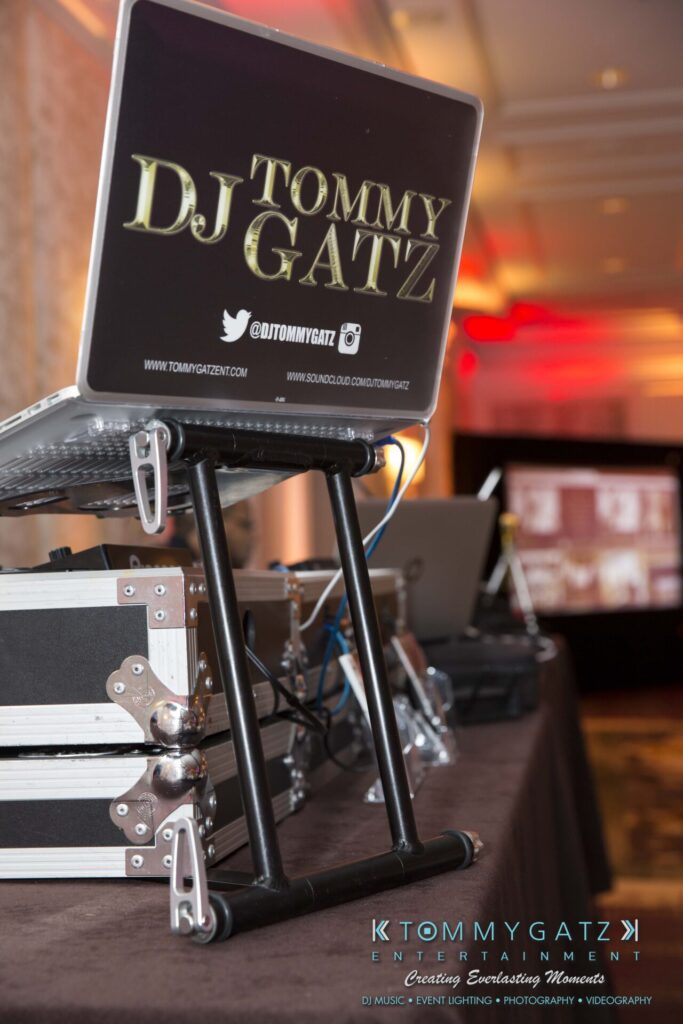 Pillar-Awards-Final-3-24-17-Blog-WM-27 - TOMMY GATZ ENTERTAINMENT A DJ setup with a laptop displaying "Tommy DJ Gatz" on the screen, next to audio equipment and cases in a dimly lit room.
