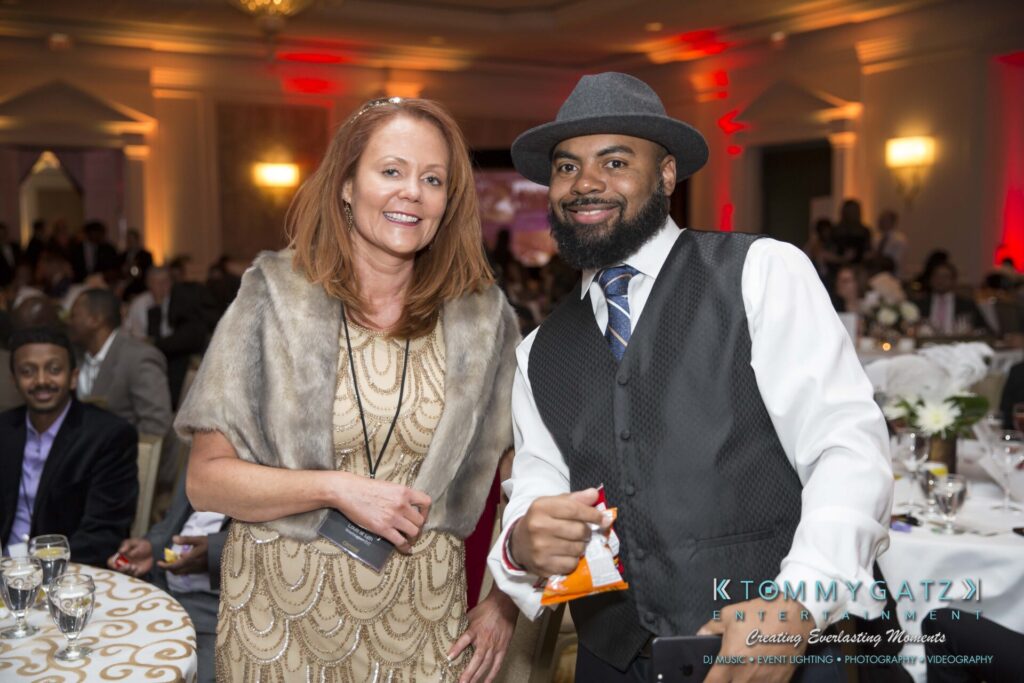 Pillar-Awards-Final-3-24-17-Blog-WM-310 - TOMMY GATZ ENTERTAINMENT A woman and a man smiling at an indoor event, surrounded by people. The man holds a small bag.