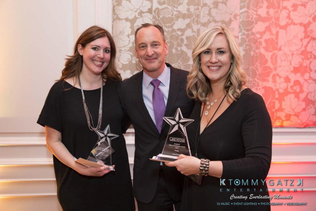 Pillar-Awards-Final-3-24-17-Blog-WM-353 - TOMMY GATZ ENTERTAINMENT Three people standing indoors, two women holding star-shaped awards and smiling, with a man in a suit in the center. They are in a decorated event space.