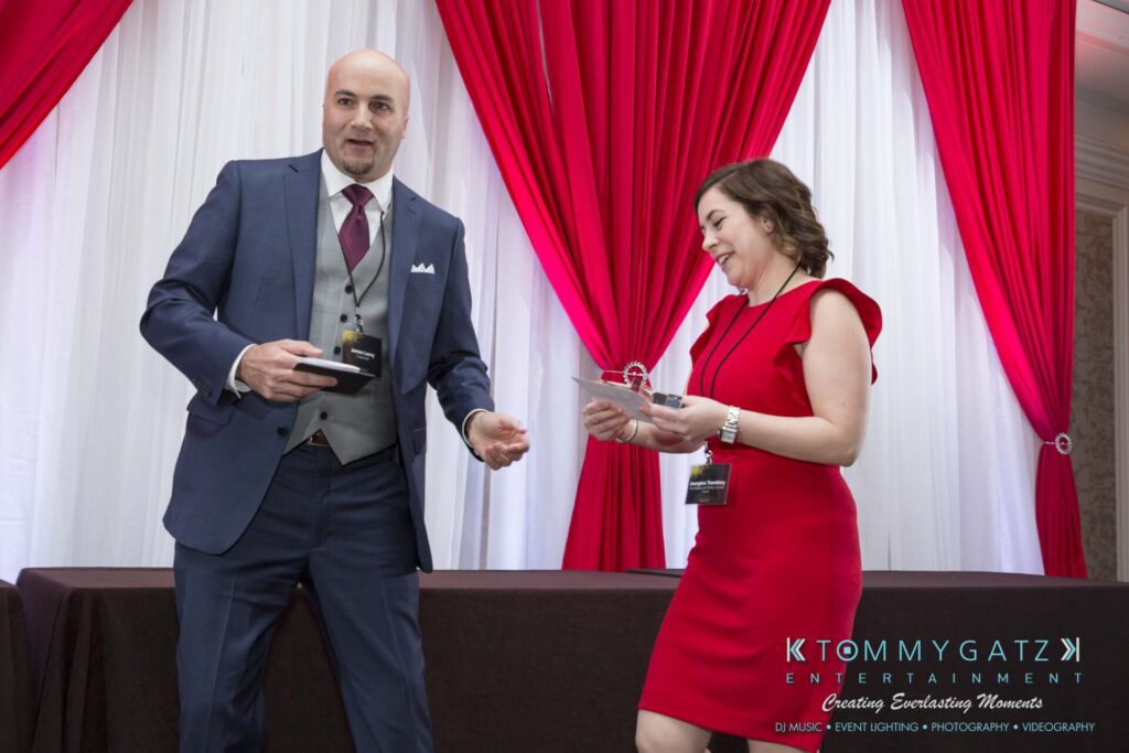 Pillar-Awards-Final-3-24-17-Blog-WM-378 - TOMMY GATZ ENTERTAINMENT A man in a suit and a woman in a red dress stand talking in front of red and white drapes, with a table behind them. They are holding papers and wearing name badges.