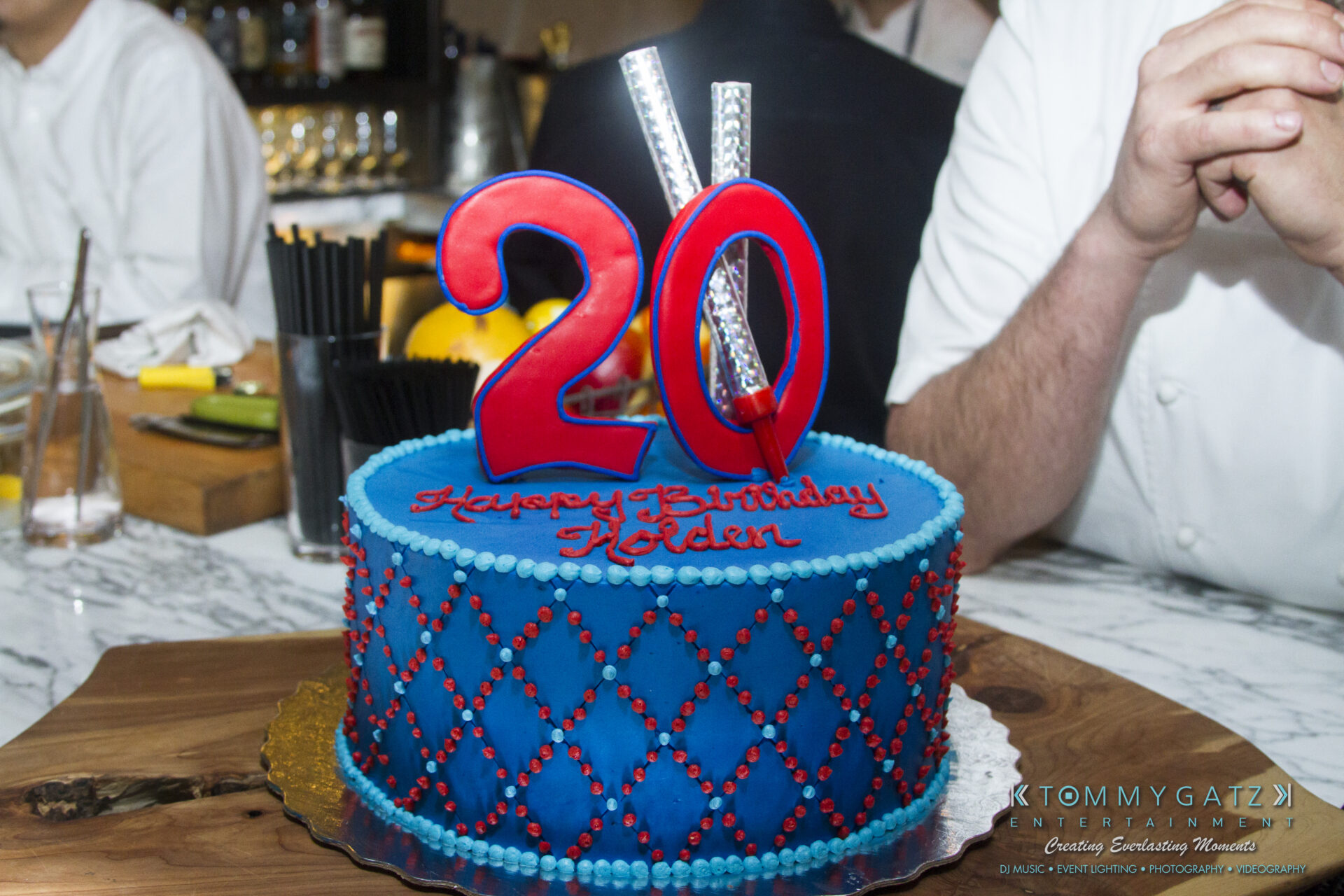 SUSAN_50TH_BIRTHDAY_WATERMARK128 - TOMMY GATZ ENTERTAINMENT A blue birthday cake with red and silver decorations, featuring the number 20 on top and "Happy Birthday Eden" written on it.