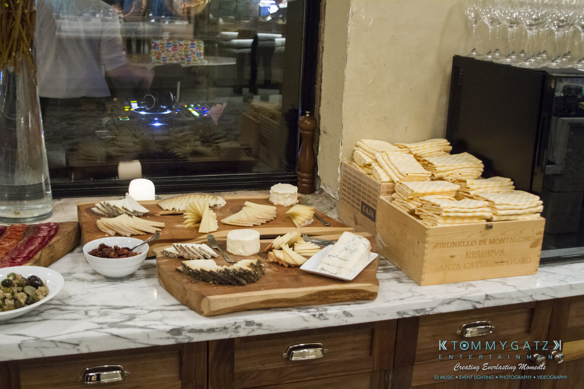 SUSAN_50TH_BIRTHDAY_WATERMARK2 - TOMMY GATZ ENTERTAINMENT A variety of cheeses and sliced meats are arranged on wooden boards, accompanied by a bowl of olives and a box of breadsticks, set on a marble countertop.