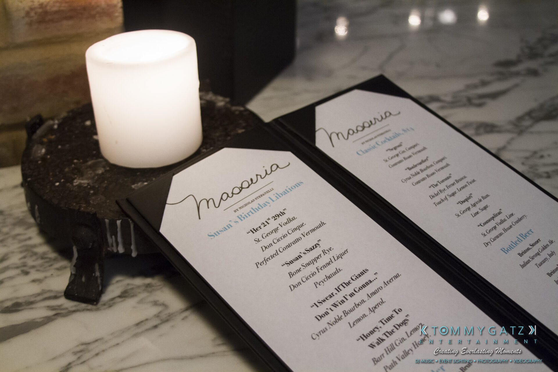 SUSAN_50TH_BIRTHDAY_WATERMARK24 1 - TOMMY GATZ ENTERTAINMENT A candle on a stone holder next to two black-bordered cocktail menus on a marble table.