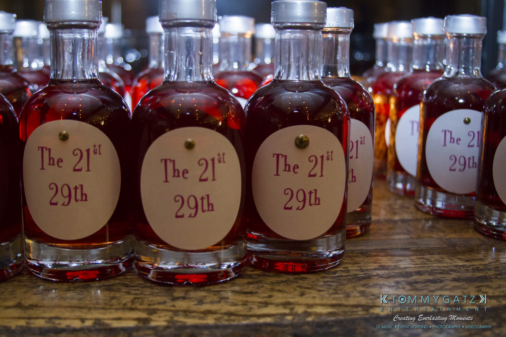 SUSAN_50TH_BIRTHDAY_WATERMARK30 1 - TOMMY GATZ ENTERTAINMENT Clear bottles filled with a dark liquid, each labeled "The 21st 29th," are arranged on a wooden surface.