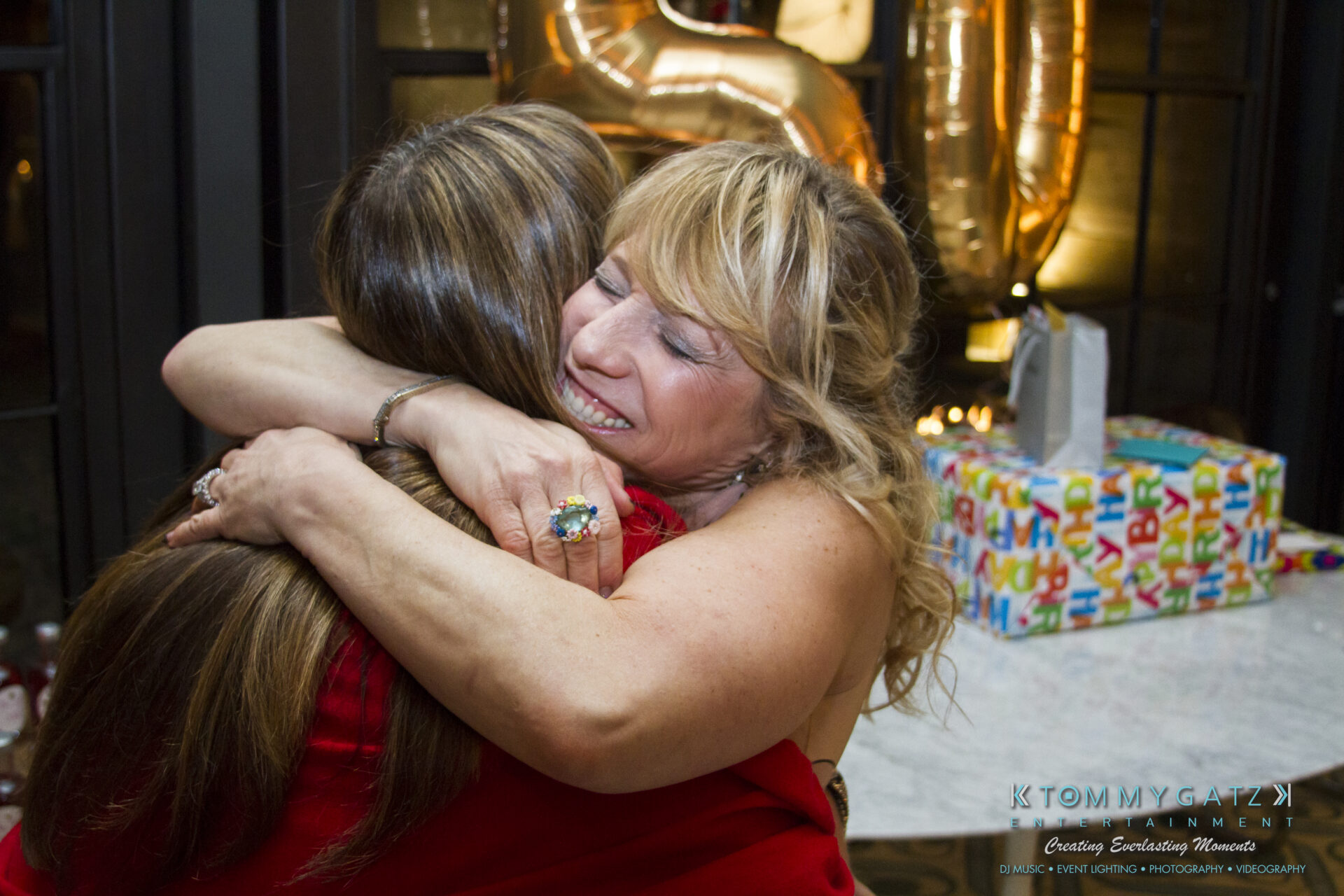 SUSAN_50TH_BIRTHDAY_WATERMARK31 - TOMMY GATZ ENTERTAINMENT Two people embrace warmly in front of large gold balloons and a stack of gift-wrapped presents on a table.
