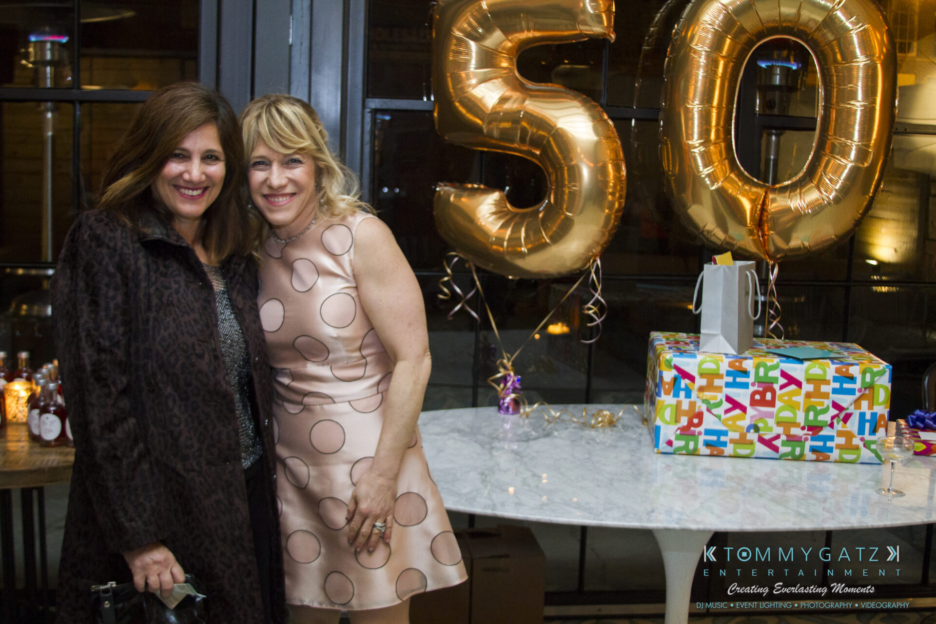 SUSAN_50TH_BIRTHDAY_WATERMARK33 - TOMMY GATZ ENTERTAINMENT Two women posing and smiling in front of large gold "50" balloons at a celebration, with a gift-wrapped box on a table nearby.