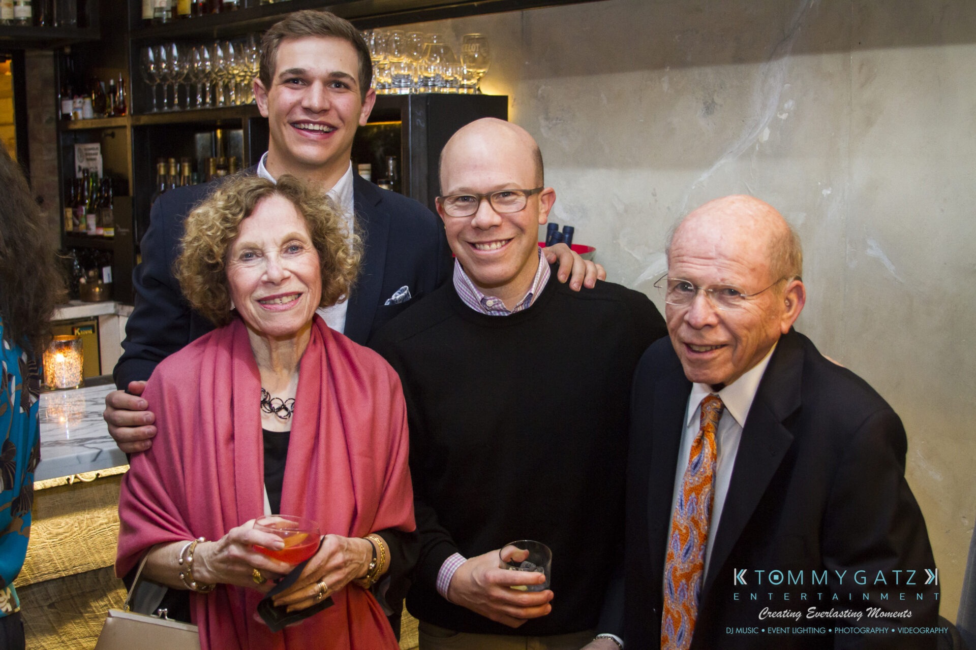 SUSAN_50TH_BIRTHDAY_WATERMARK47 - TOMMY GATZ ENTERTAINMENT Four people are gathered in a warmly lit room, smiling for the camera. The group includes two men standing behind two seniors, one woman and one man. They each hold a drink.