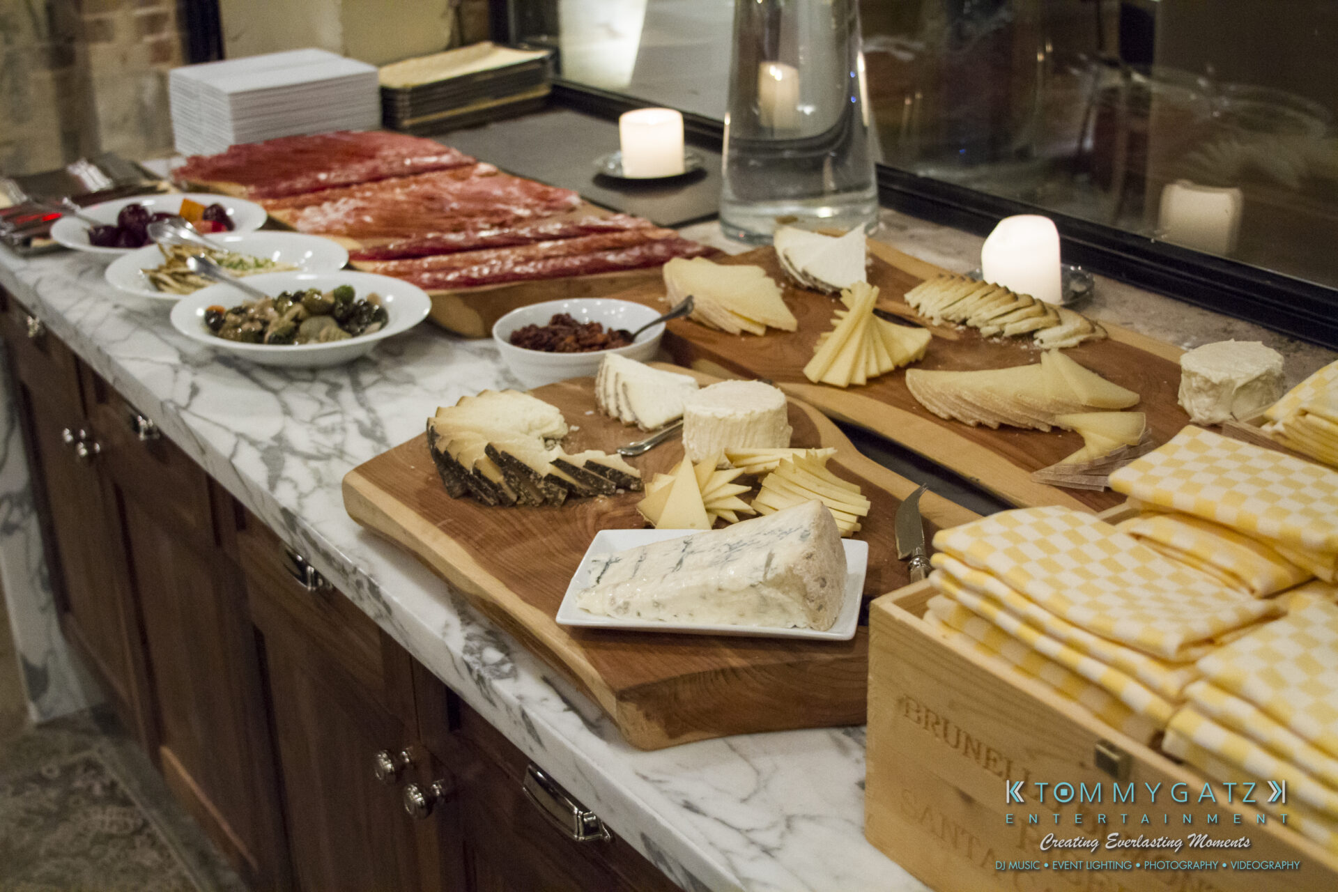 SUSAN_50TH_BIRTHDAY_WATERMARK5 - TOMMY GATZ ENTERTAINMENT A marble countertop displays a selection of cheese, meats, and olives, accompanied by napkins and a candle.