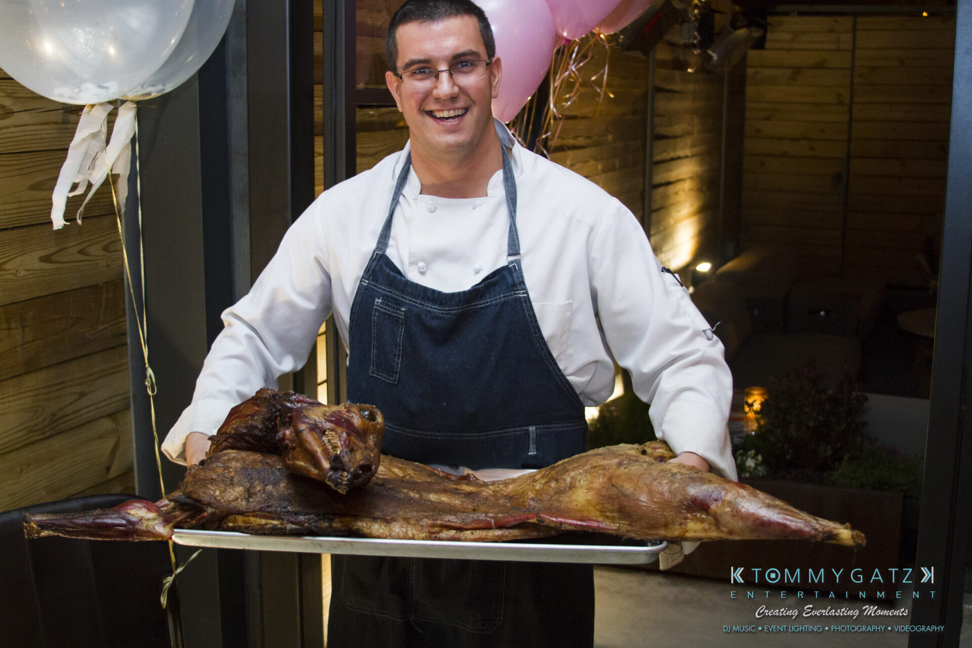 SUSAN_50TH_BIRTHDAY_WATERMARK60 - TOMMY GATZ ENTERTAINMENT Chef holding a tray with a roasted whole animal, smiling, standing indoors. Balloons visible in the background.