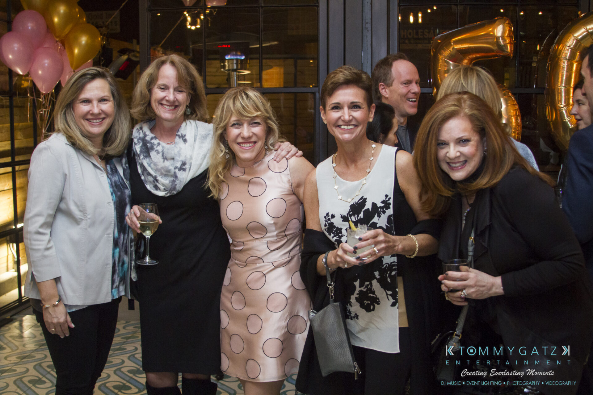 SUSAN_50TH_BIRTHDAY_WATERMARK63 - TOMMY GATZ ENTERTAINMENT A group of women standing together at a party, smiling and holding drinks. Balloon decorations are visible in the background.