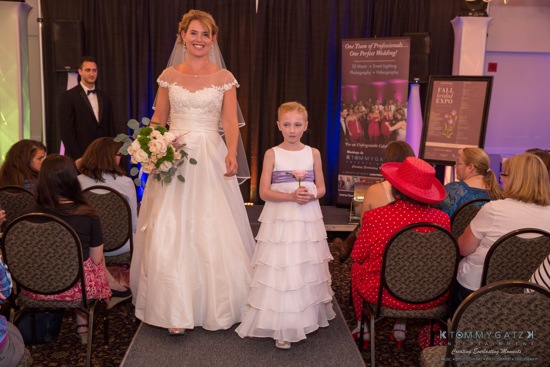 untitled-10 - TOMMY GATZ ENTERTAINMENT A bride and a young girl in white dresses walk down an aisle at a bridal event. Audience members sit on either side.