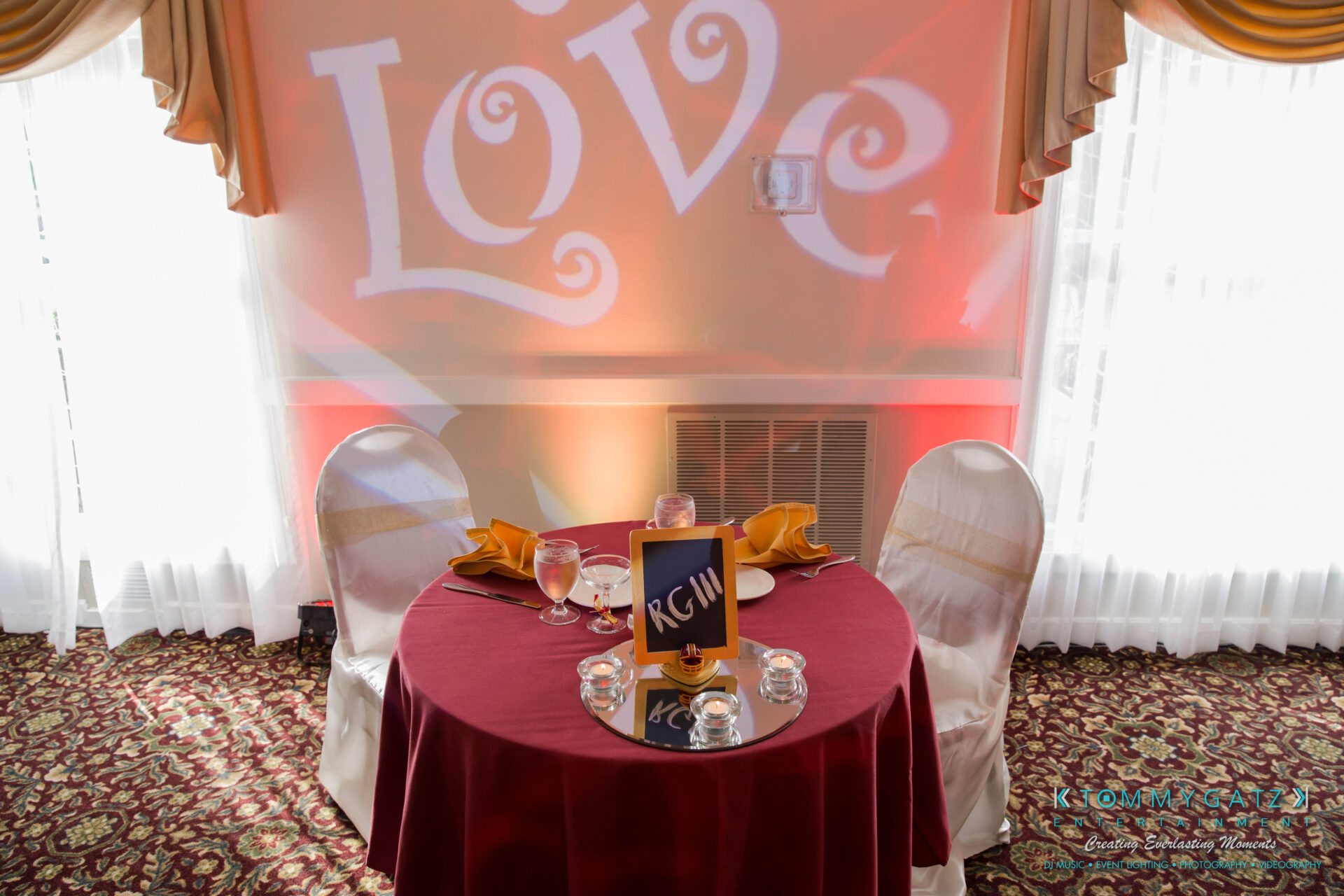 untitled-12 - TOMMY GATZ ENTERTAINMENT A round table with a red tablecloth is set for two, featuring yellow napkins, water glasses, and a menu card labeled "RCW." The backdrop displays the word "Love" illuminated in orange.