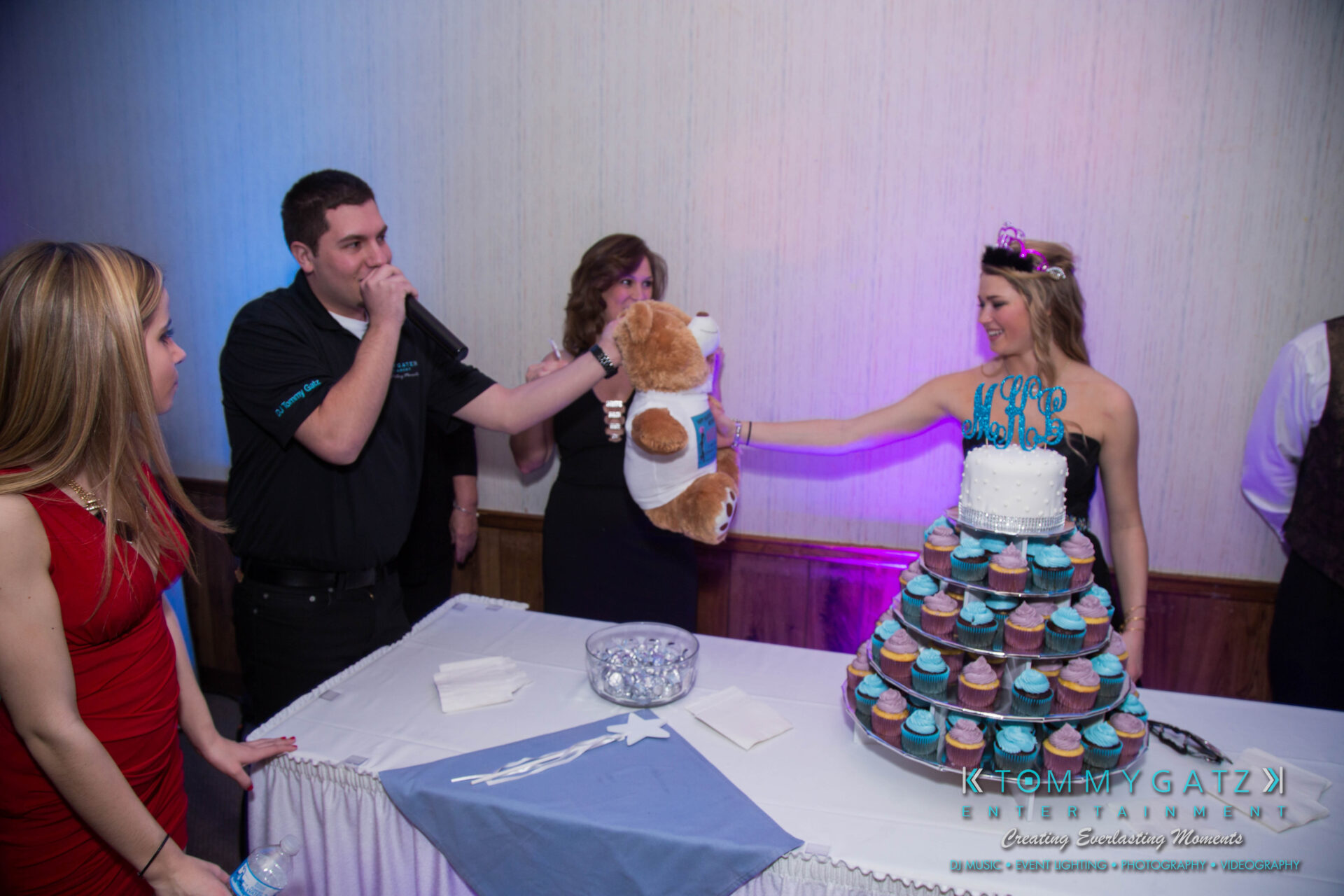 untitled-15 1 - TOMMY GATZ ENTERTAINMENT A woman in a blue dress holds a stuffed animal across a table with a cupcake tower. A man with a microphone stands nearby.