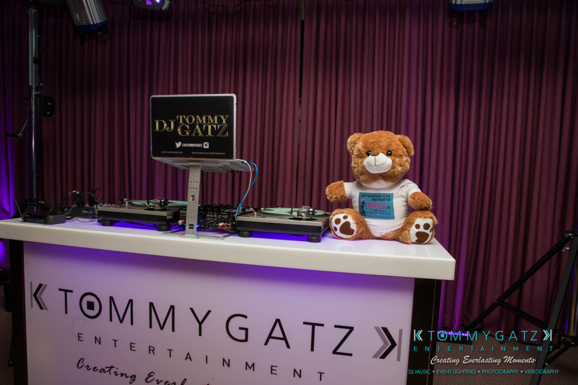untitled-18 - TOMMY GATZ ENTERTAINMENT DJ booth with Tommy Gatz Entertainment branding, featuring turntables and a teddy bear sitting beside them.