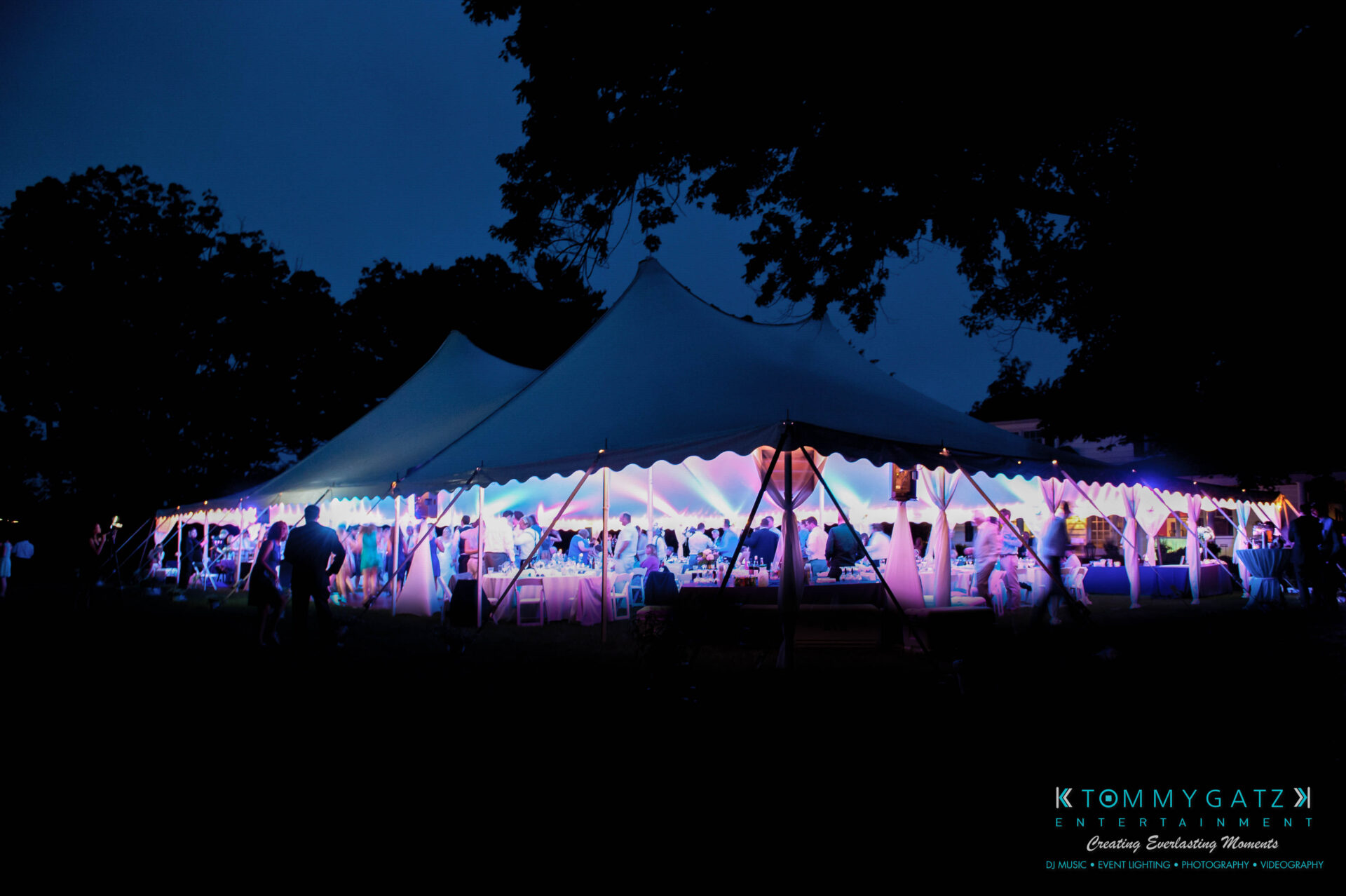 untitled-26 - TOMMY GATZ ENTERTAINMENT A large tent illuminated with colorful lights stands outdoors at dusk, surrounded by silhouetted trees and people walking nearby.