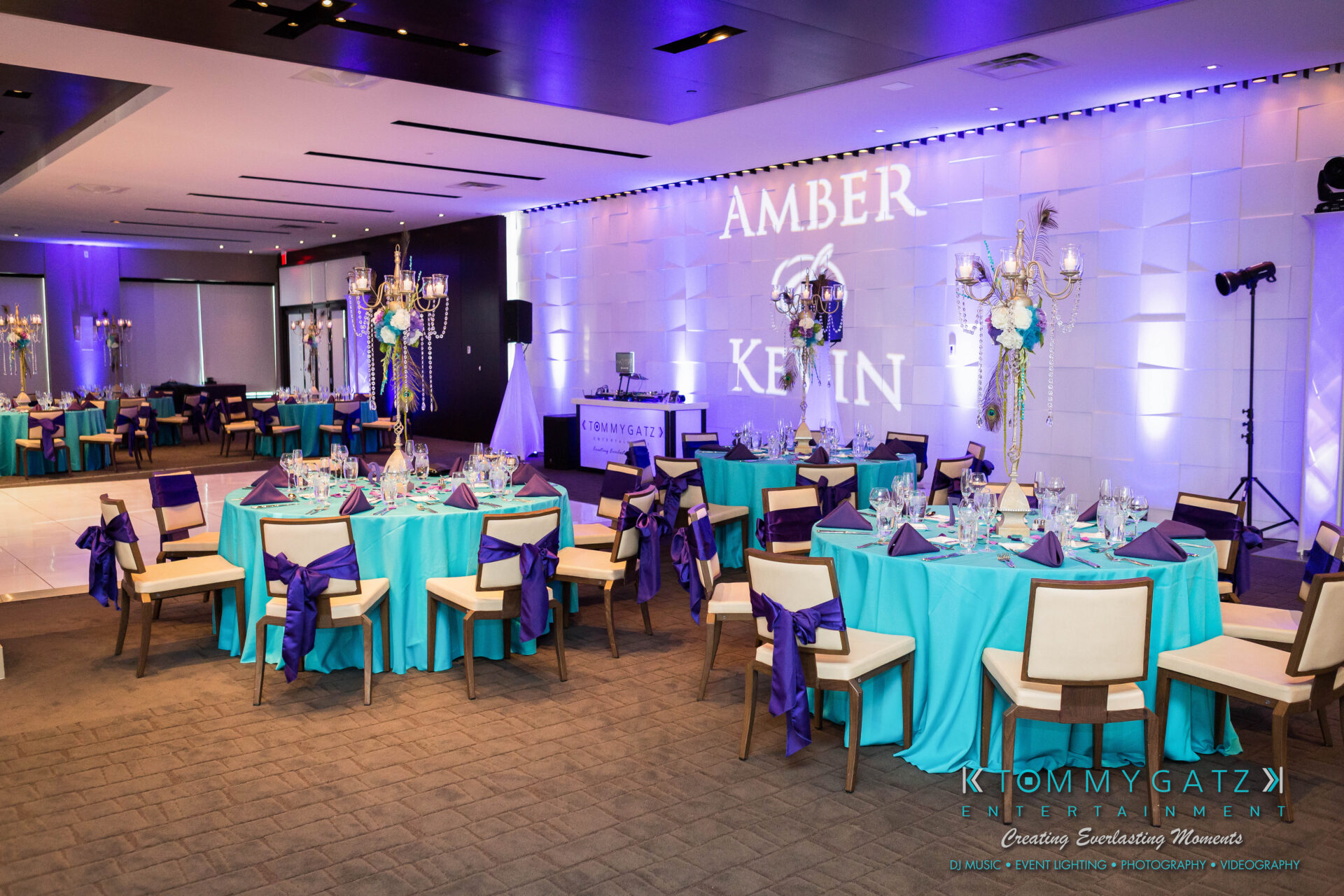 untitled-27 - TOMMY GATZ ENTERTAINMENT Elegant wedding reception setup with turquoise and purple decorations, round tables, ornate centerpieces, and a wall projection displaying "Amber & Kevin.