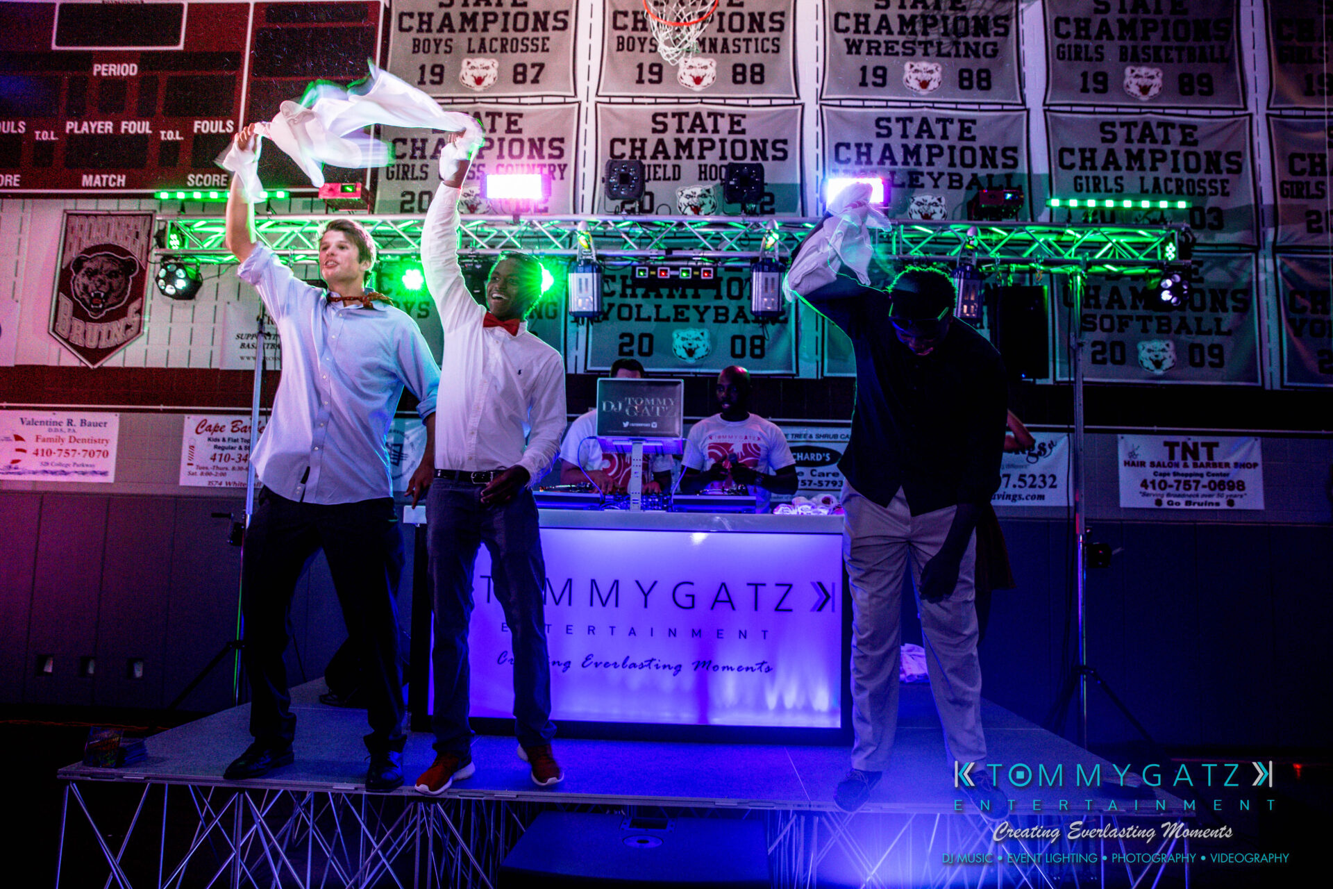 untitled-28 - TOMMY GATZ ENTERTAINMENT Three people on a stage raise their hands, holding napkins, in front of a DJ booth with colorful lights and a banner displaying "Tommy Gatz Entertainment.