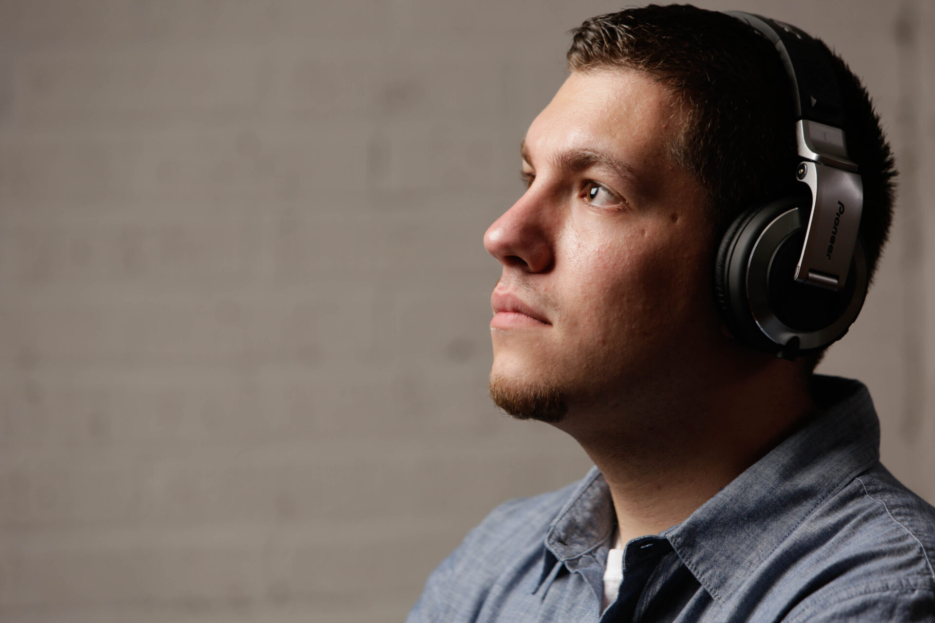 untitled-30 - TOMMY GATZ ENTERTAINMENT A man in a blue shirt is wearing headphones and looking upward, against a blurred gray brick background.