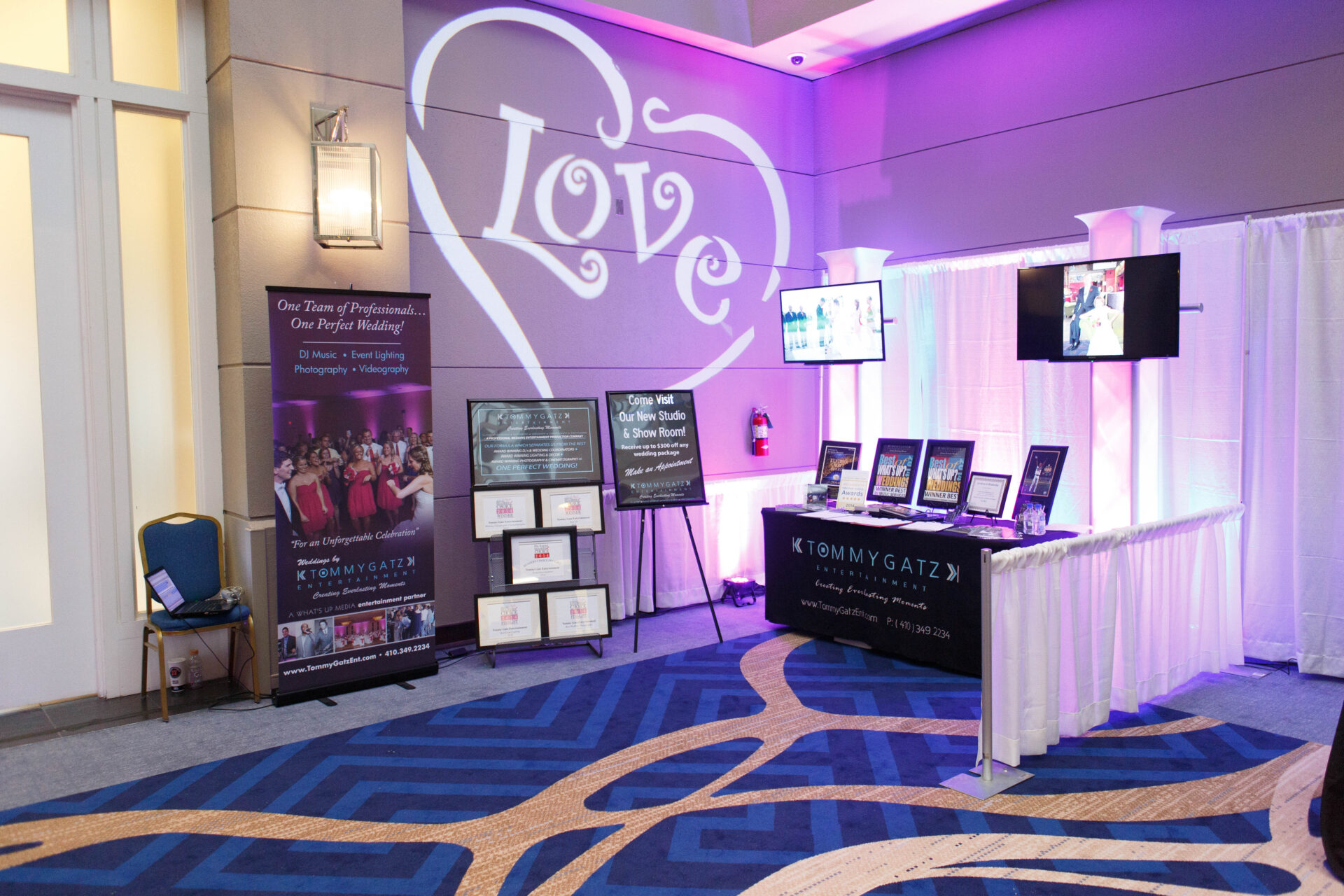 untitled-31 - TOMMY GATZ ENTERTAINMENT Wedding event booth displaying banners, framed photos, and screens under purple lighting, with a "Love" projection on the wall.