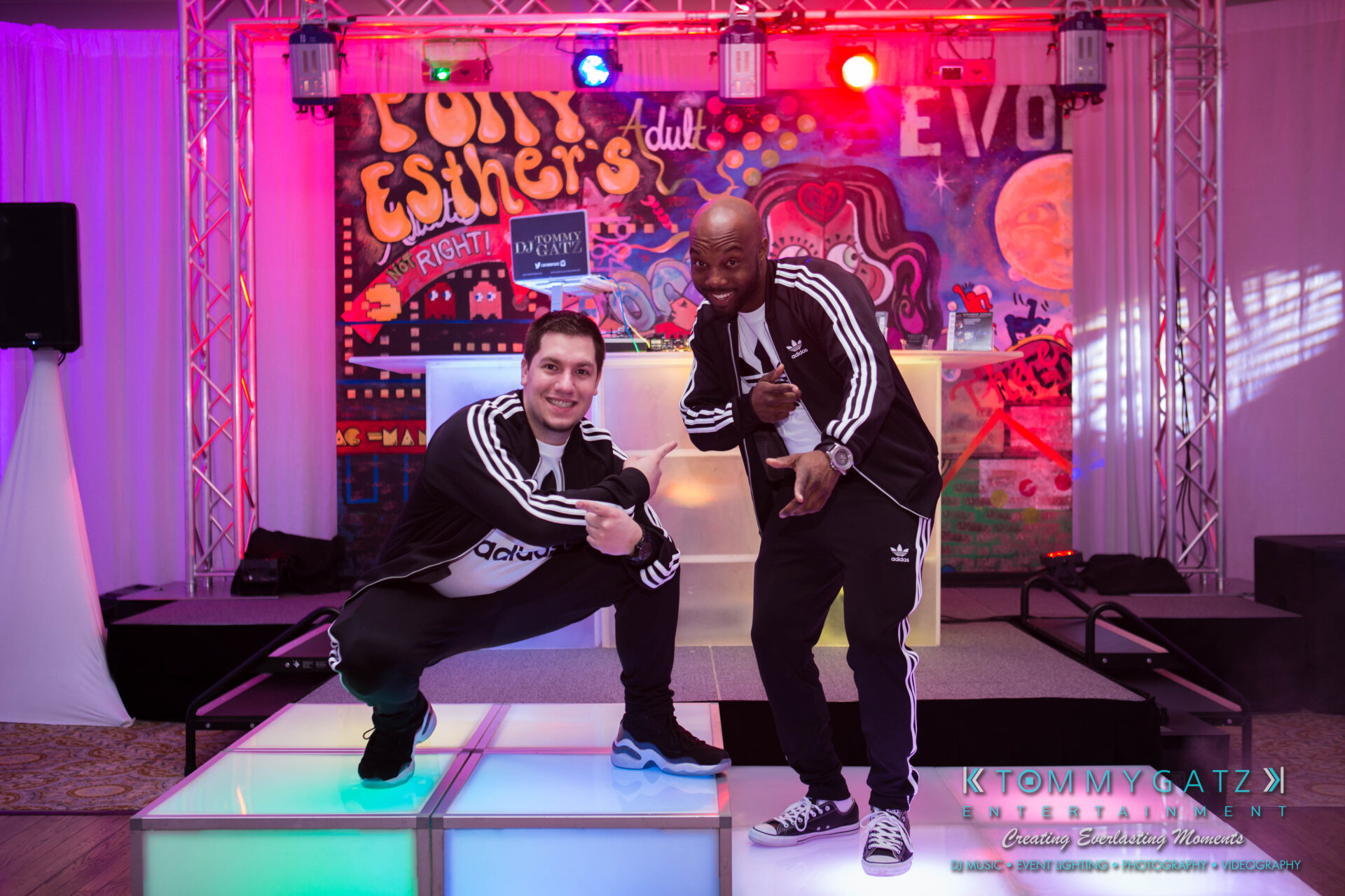 untitled-4 - TOMMY GATZ ENTERTAINMENT Two people in Adidas tracksuits pose on a lit-up platform with a colorful graffiti backdrop.