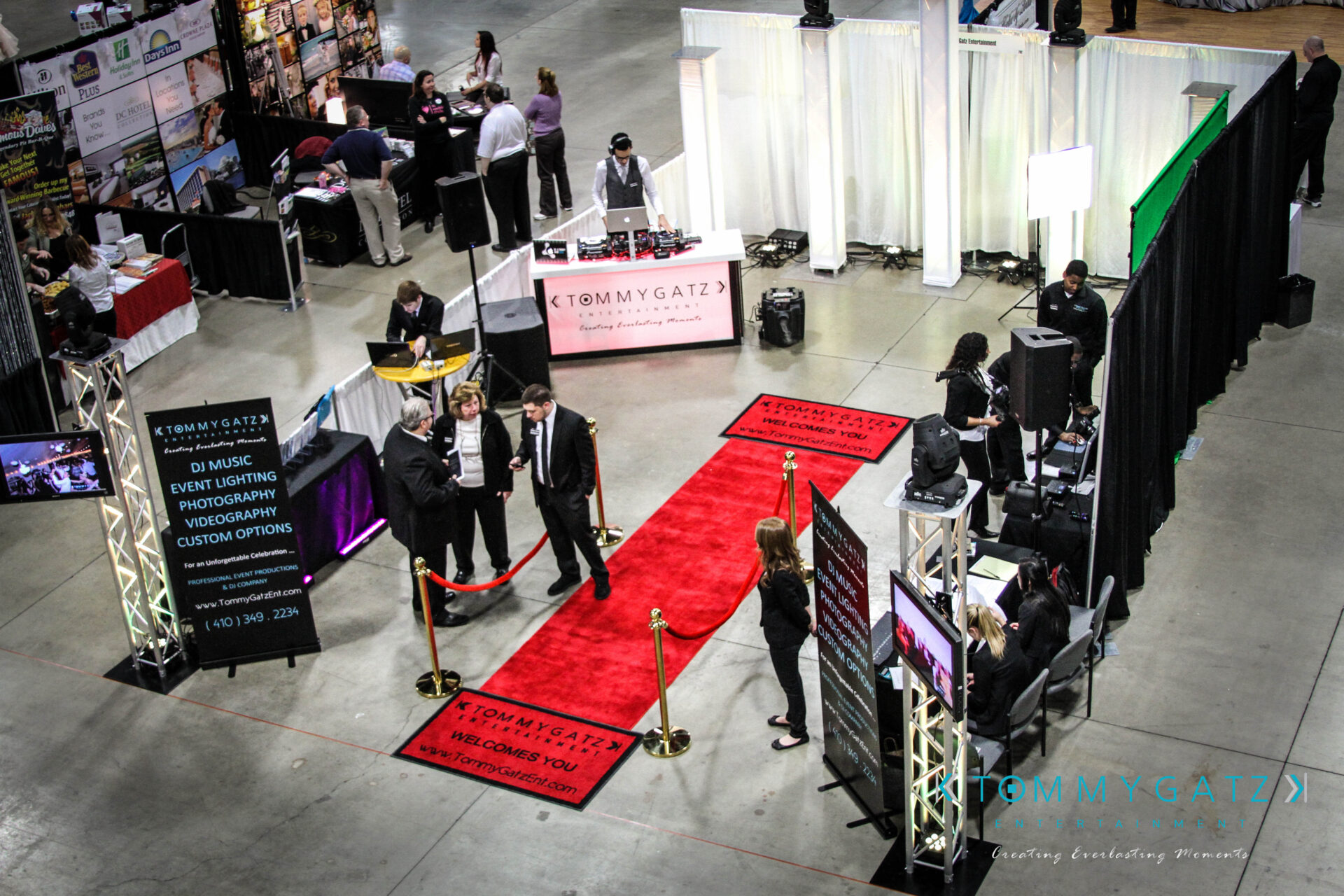 untitled-43 - TOMMY GATZ ENTERTAINMENT Trade show with a red carpet and Tommy Gatz branding. Attendees in business attire converse near booths displaying music, photography, and event services.