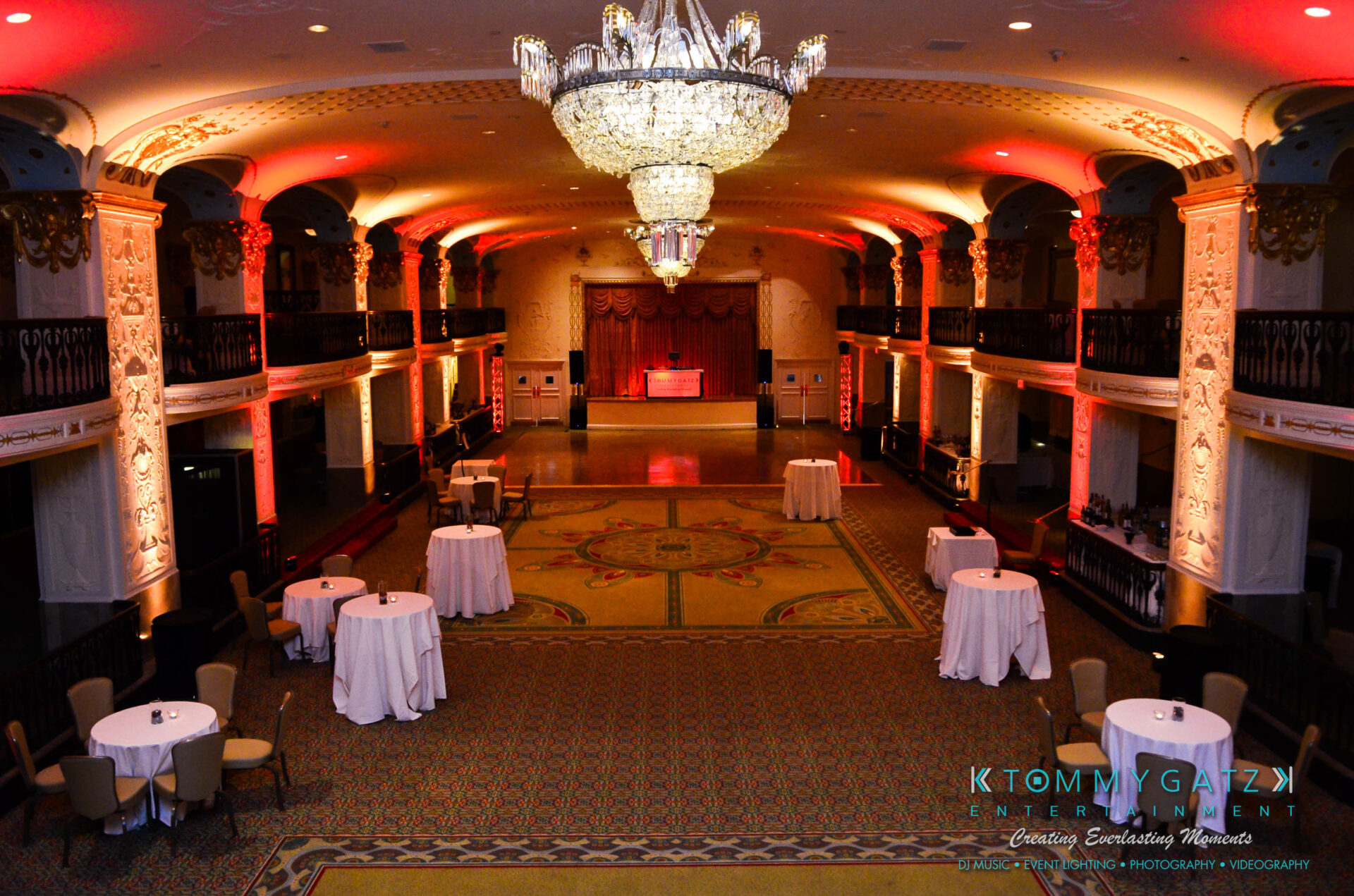 untitled-5 - TOMMY GATZ ENTERTAINMENT Elegant ballroom with round tables, white tablecloths, and a grand chandelier. Warm lighting enhances architectural details, with a stage and red curtains at the far end.