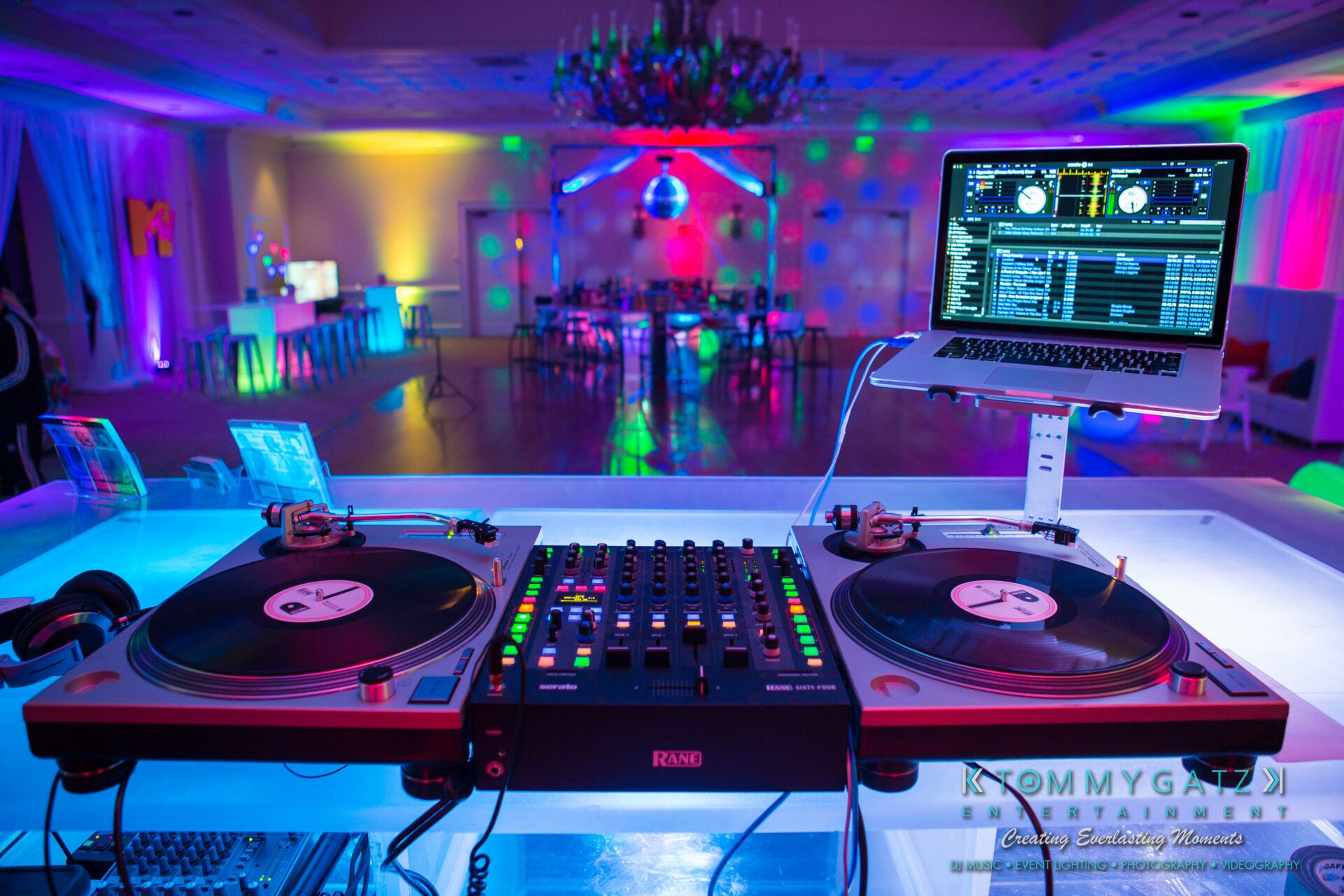 untitled-6 - TOMMY GATZ ENTERTAINMENT DJ booth with turntables and a laptop set up in a colorful, dimly lit event space, featuring a dance floor and tables in the background.