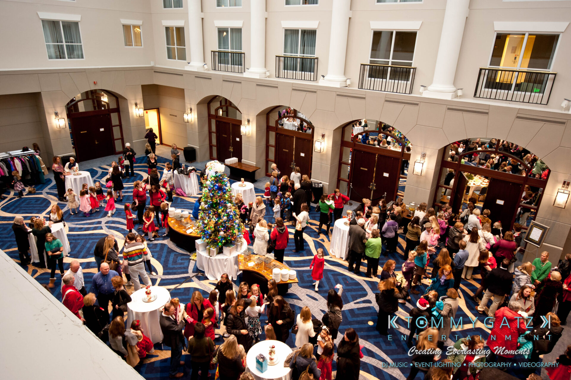 untitled-60 - TOMMY GATZ ENTERTAINMENT A large group of people mingles in a hotel lobby during a festive event with a decorated Christmas tree and banquet tables.