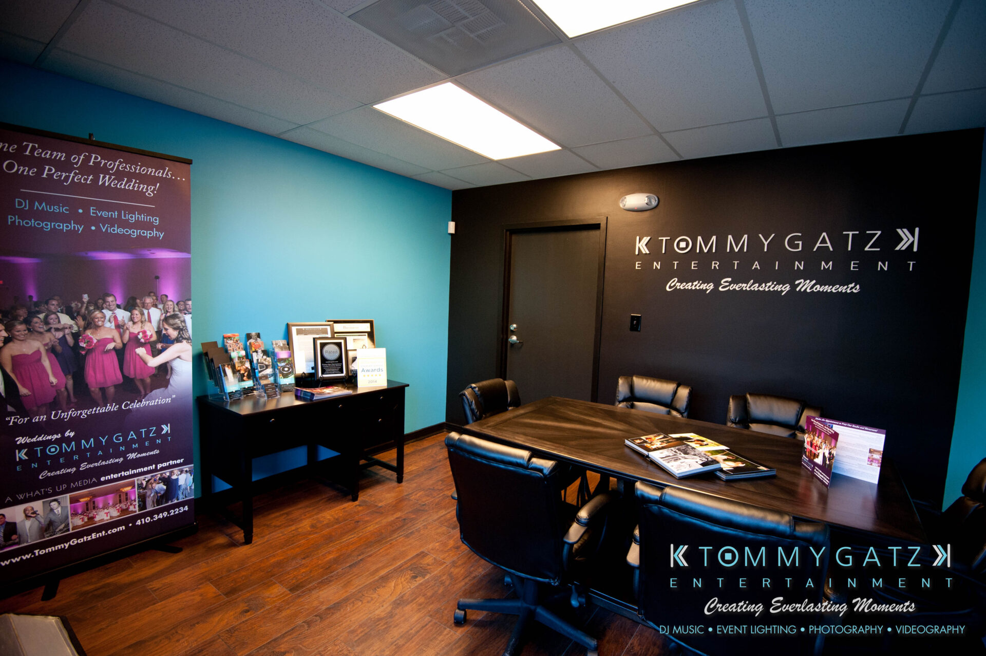 untitled-62 - TOMMY GATZ ENTERTAINMENT Office meeting room with a dark wood table, several chairs, and a promotional banner for Tommy Gatz Entertainment on the left. Walls are painted blue and white.