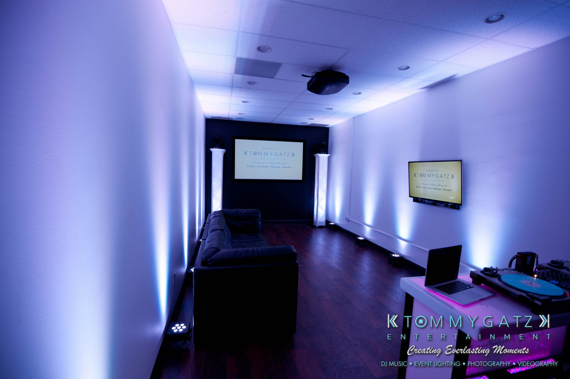 untitled-63 - TOMMY GATZ ENTERTAINMENT Modern room with a couch, DJ table, and two screens displaying "Tommy Gatz Entertainment." Ceiling lights and illuminated columns enhance the ambiance.