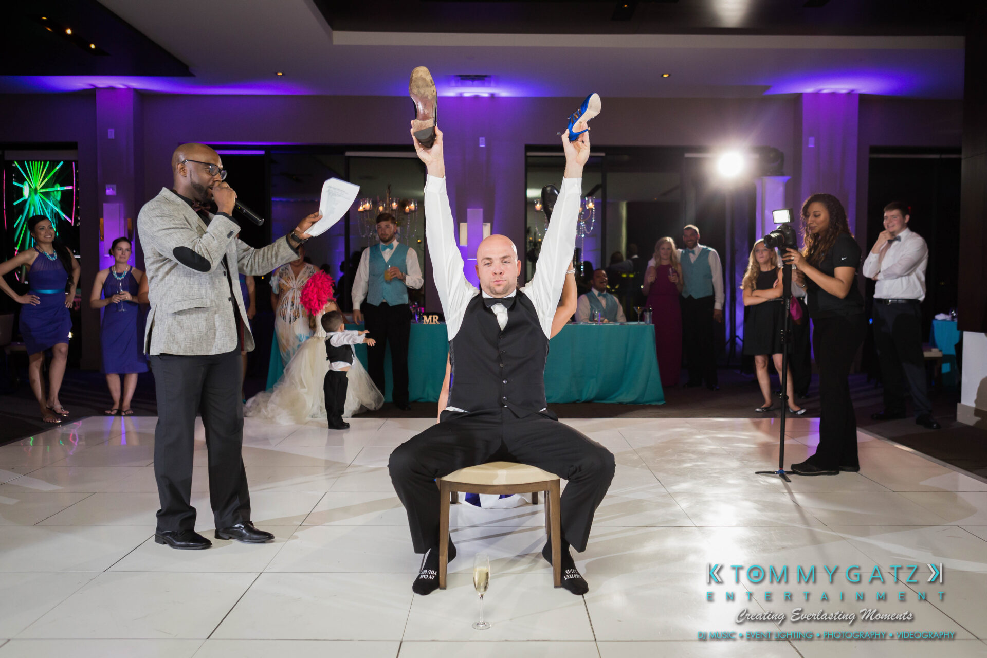 untitled-7 - TOMMY GATZ ENTERTAINMENT Man sitting on a chair at a wedding, holding up two shoes, facing an audience. Host with a microphone stands nearby. Guests watch and smile in the background.
