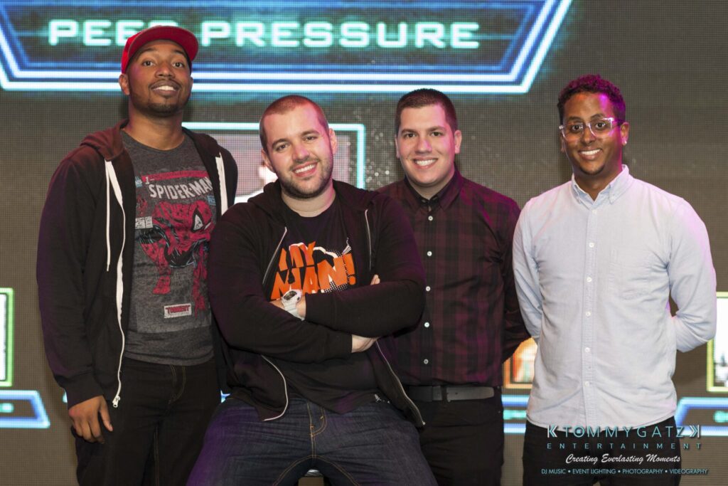 18A0008 - TOMMY GATZ ENTERTAINMENT Four men standing and posing for a photo indoors, smiling, with a sign that reads "Peer Pressure" in the background.