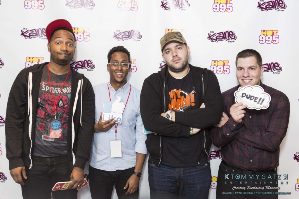 18A0020 - TOMMY GATZ ENTERTAINMENT Four people pose in front of a backdrop with "Hot 99.5" logos, holding props and smiling.