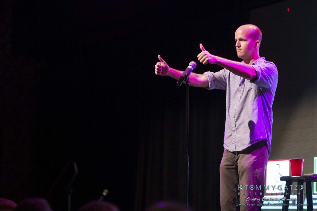 18A0031A-1 - TOMMY GATZ ENTERTAINMENT A man stands on stage performing with a microphone, giving two thumbs up.