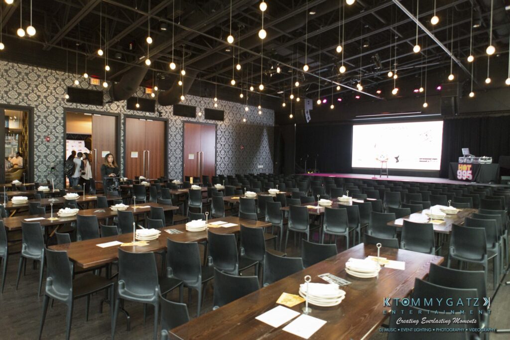 18A0038 - TOMMY GATZ ENTERTAINMENT A dimly lit event space with tables set for a meal, plates and utensils neatly arranged. Rows of chairs face a stage with a large screen displaying white and purple graphics.