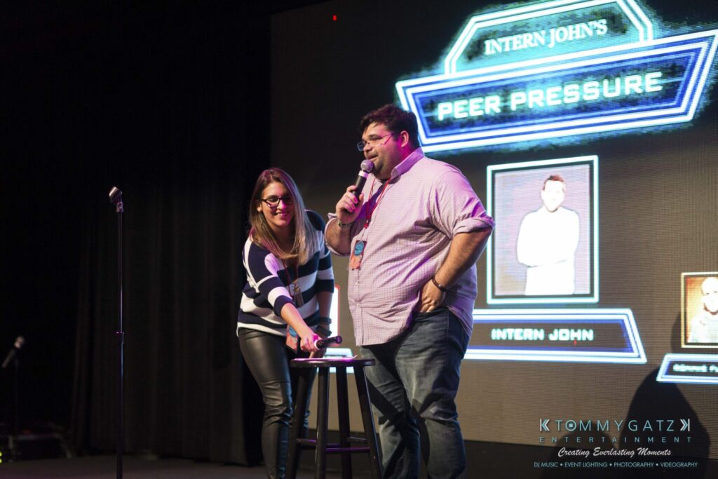18A0042A-1 - TOMMY GATZ ENTERTAINMENT Two people stand on stage with microphones in front of a sign that reads "Intern John's Peer Pressure." They are near a small round table.