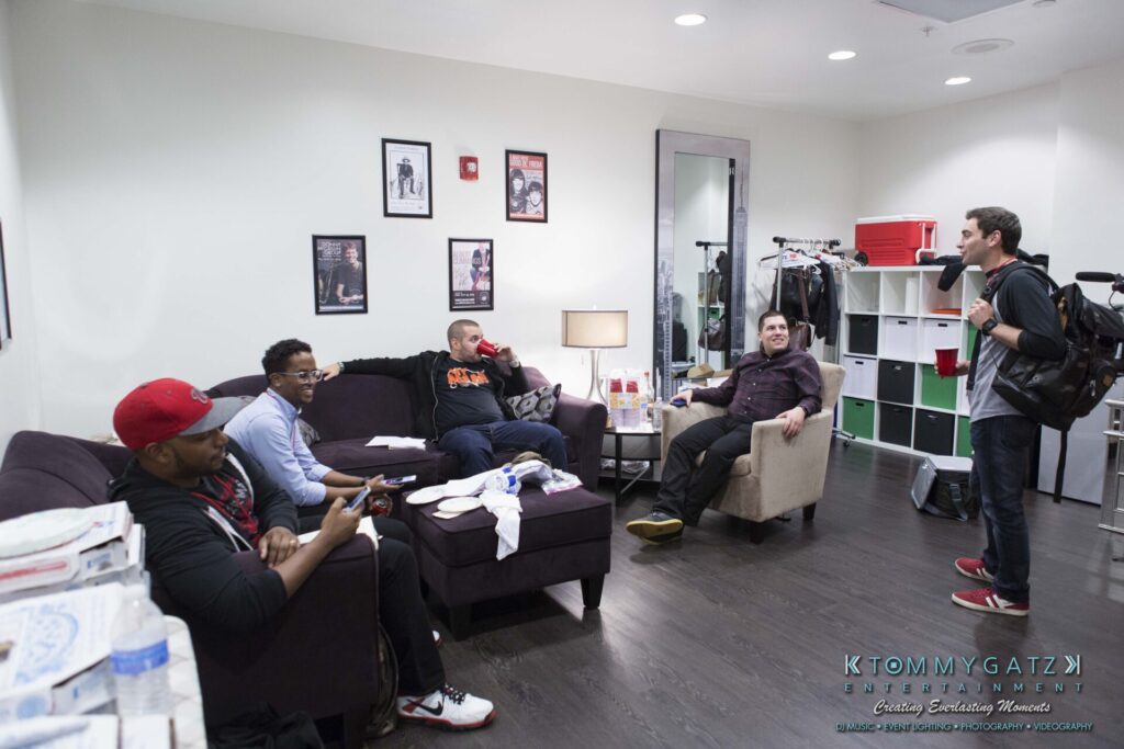 18A0062 - TOMMY GATZ ENTERTAINMENT A group of people casually sitting and talking in a living room, with a mix of couches and chairs, papers, and a pizza box visible.