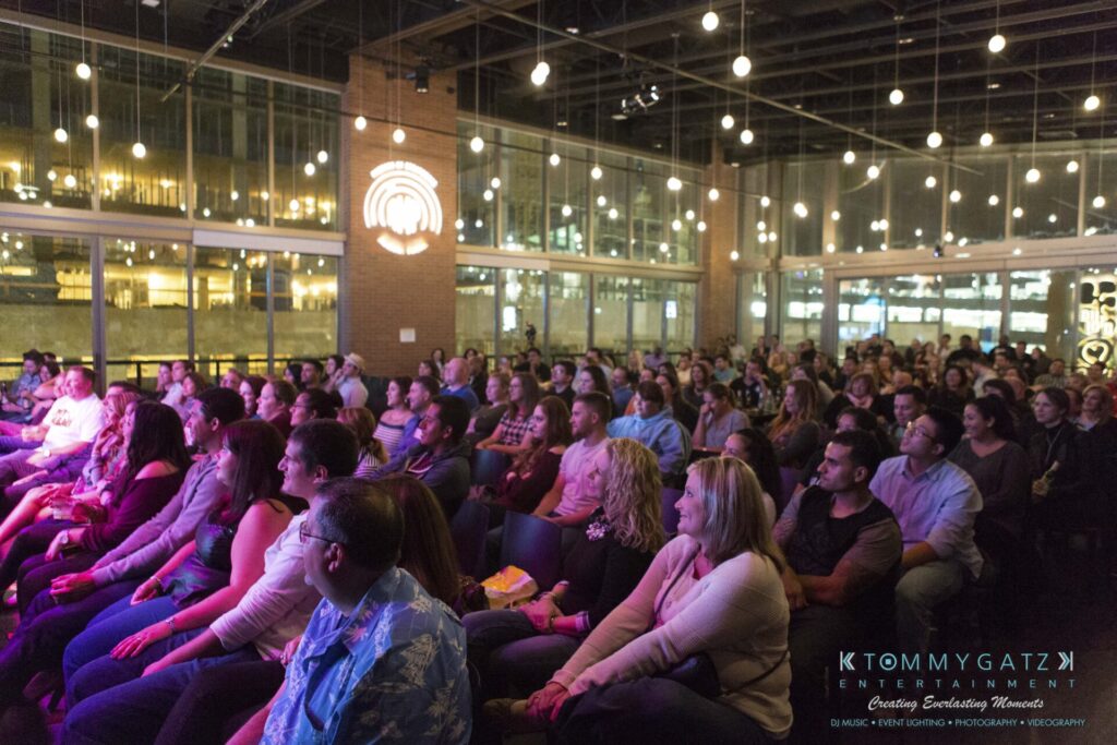 18A0122A - TOMMY GATZ ENTERTAINMENT Audience seated at an indoor event, attentively watching a stage performance, with string lights hanging from the ceiling and large windows in the background.