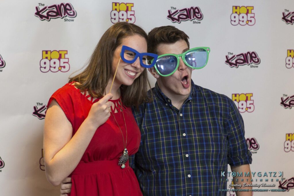 18A0131 - TOMMY GATZ ENTERTAINMENT Two people pose playfully with oversized glasses in front of a step and repeat banner featuring "The Kane Show" and "HOT 99.5" logos.