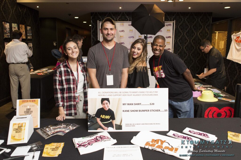 18A0135 - TOMMY GATZ ENTERTAINMENT Four people stand behind a merchandise table displaying T-shirts, stickers, and a donation sign for charity in a lobby setting.